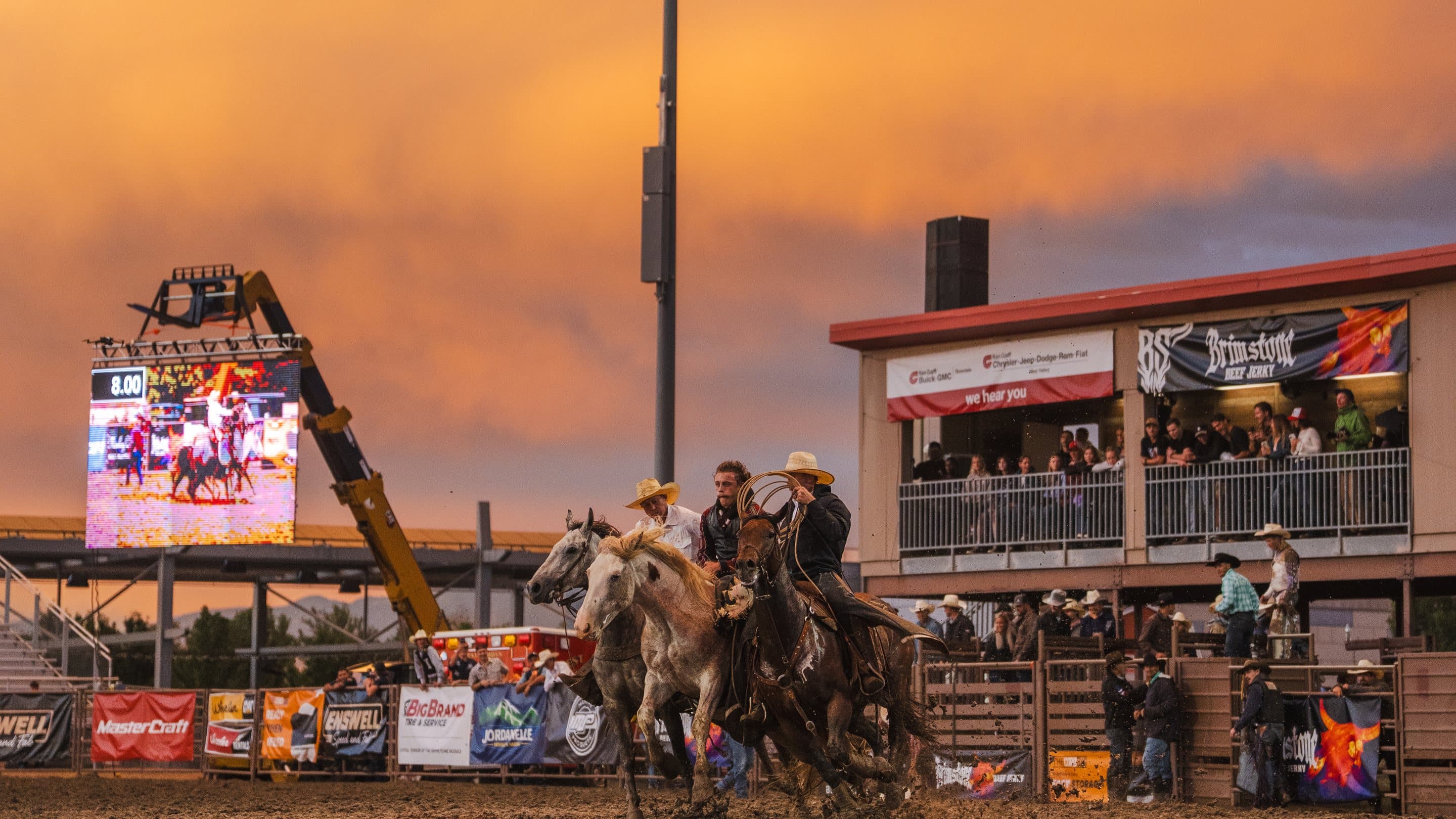Pickup riders crossing the arena at the Brimstone Rodeo under a burning sunset, the Brimstone Beef Jerky banner on the announcer's box behind them